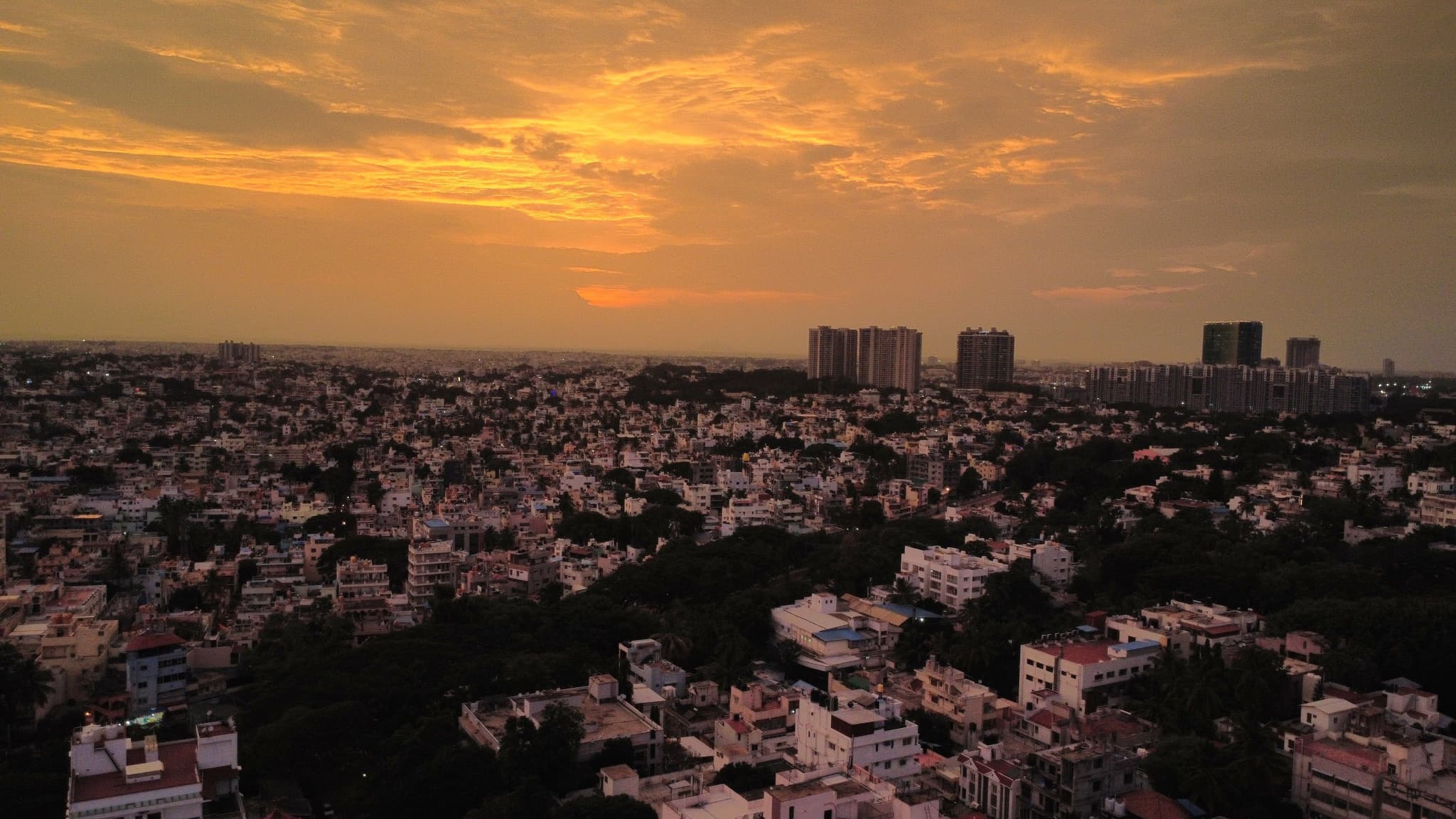 A dense cityscape stretching to the horizon under an orange sunset with distant high-rise buildings silhouetted in the background