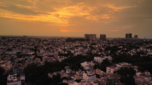 A dense cityscape stretching to the horizon under an orange sunset with distant high-rise buildings silhouetted in the background