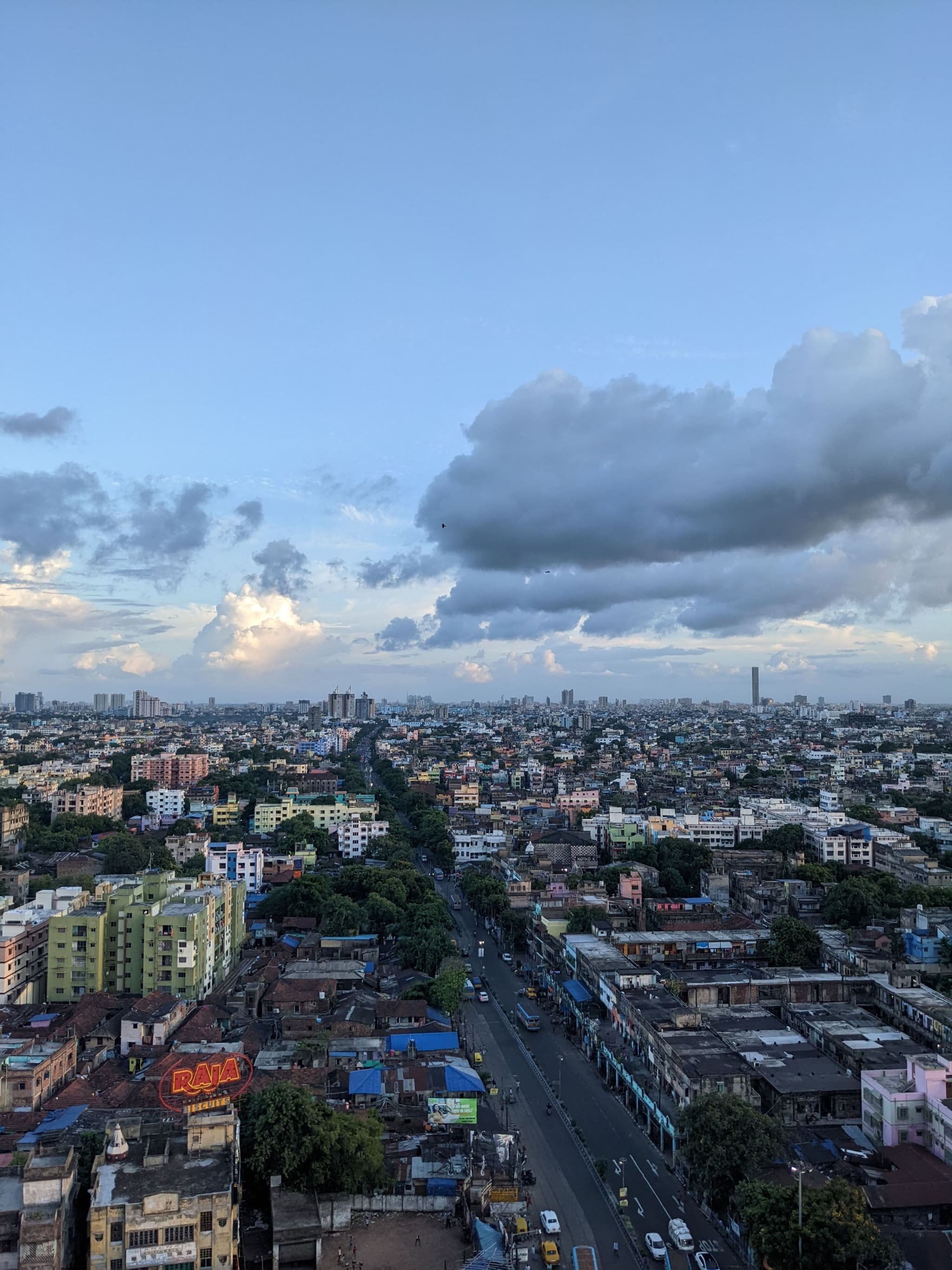 A sprawling urban landscape with a central road stretching into the distance, flanked by colorful buildings. Large clouds and a blue sky create a dramatic skyline, with high-rise buildings visible on the horizon