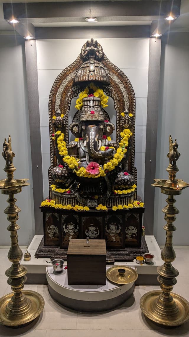 Ornate Hindu shrine with a black stone deity idol adorned in yellow and pink flower garlands, set in a white alcove and flanked by two tall brass oil lamps, with offerings placed on a curved pedestal below