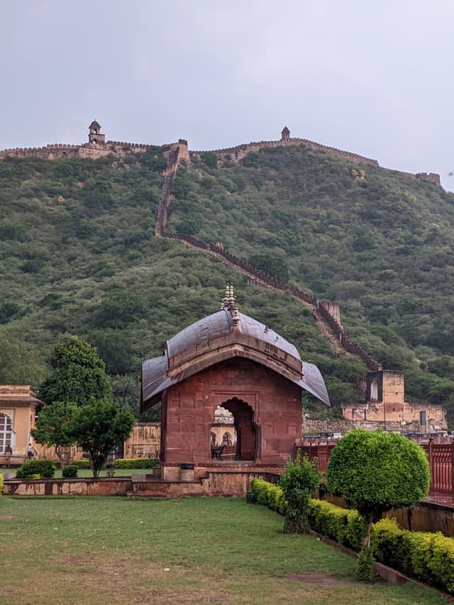A small stone archway stands on lush green grass, with an ancient fort and winding wall set against a verdant hillside under a cloudy sky