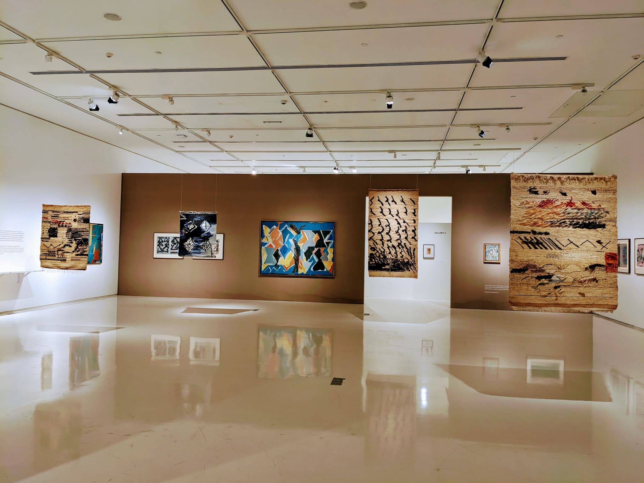 A modern art gallery with various paintings displayed on a brown feature wall and a reflective polished floor