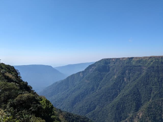 A vast mountain range under a clear blue sky, with steep cliffs and green vegetation