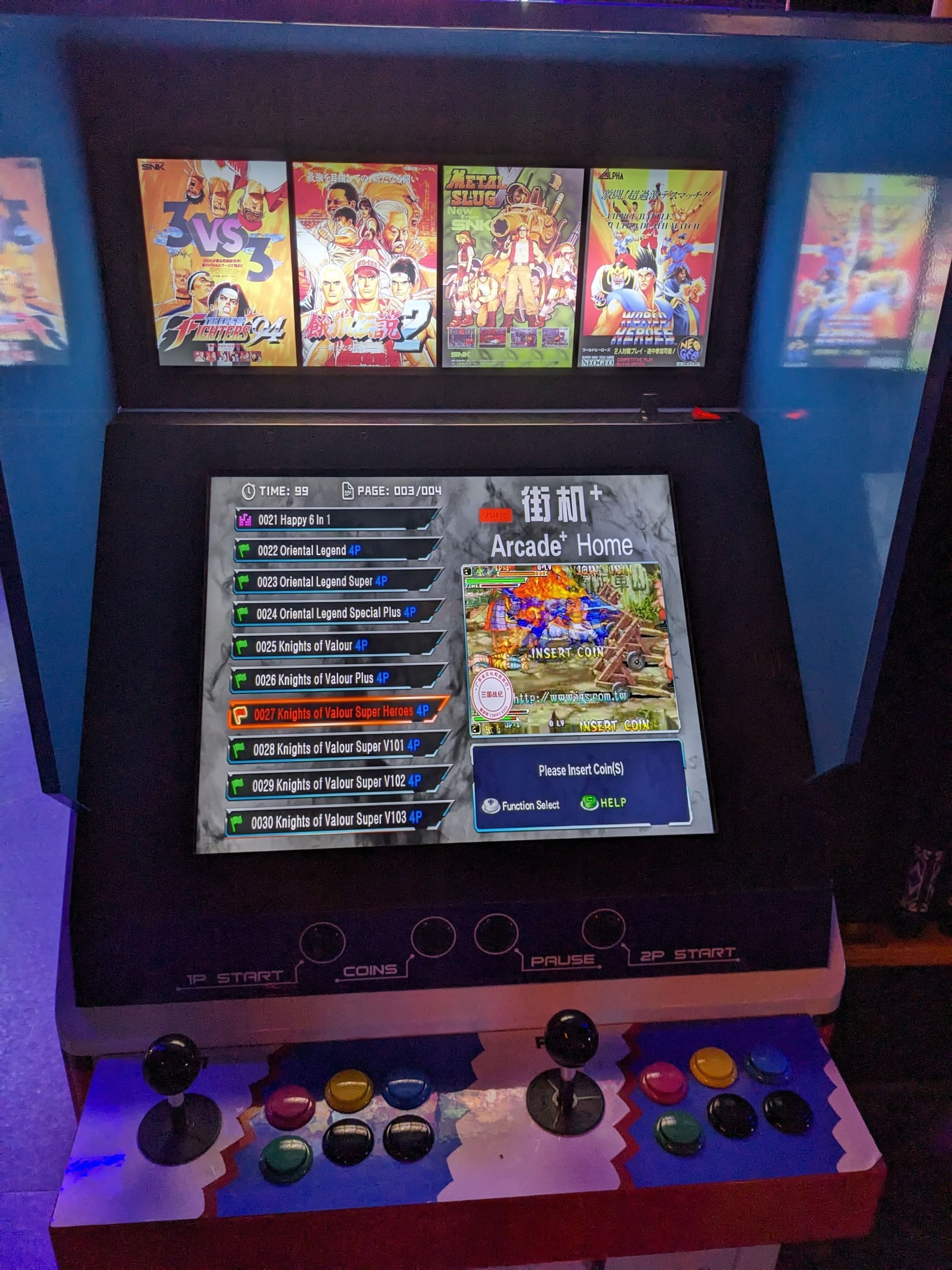 An arcade machine with a colorful control panel featuring multiple joysticks and buttons. The screen displays a menu with various game titles, and the top section shows several game covers