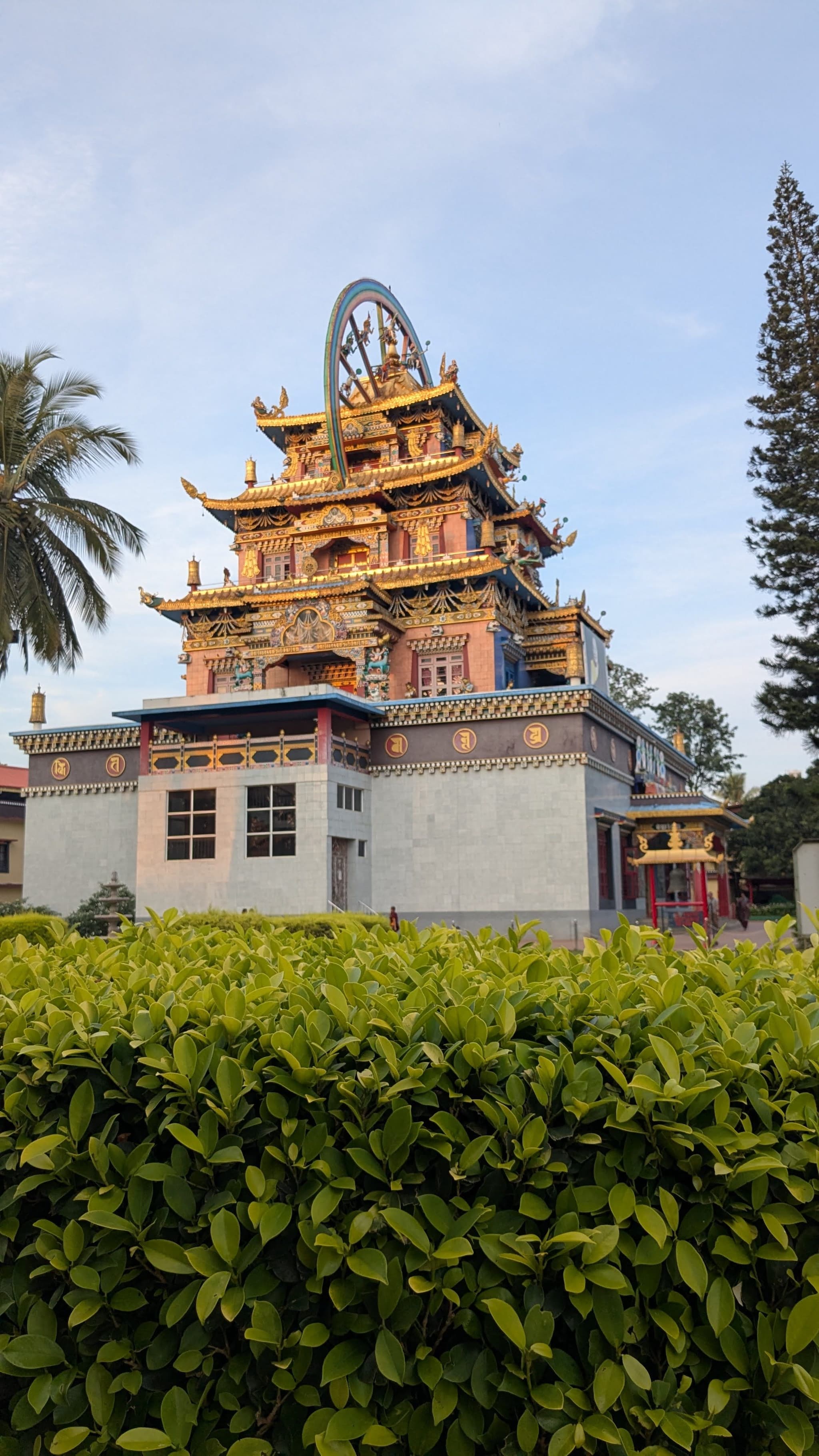 Ornate temple tower rising above a hedge, framed by palm and conifer trees under an evening sky