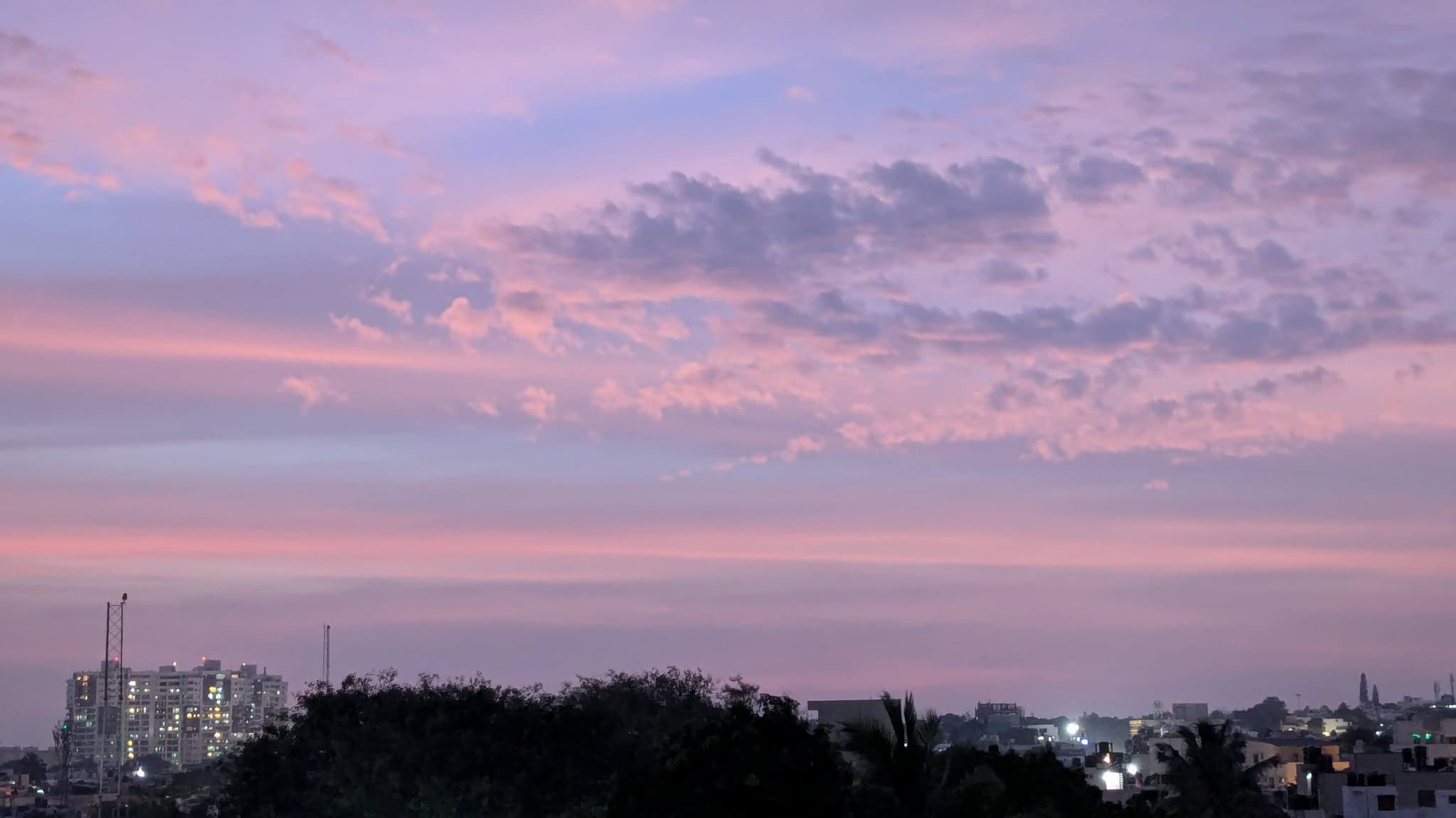 City skyline with a pink and purple sunset sky, silhouetted trees, and a construction site