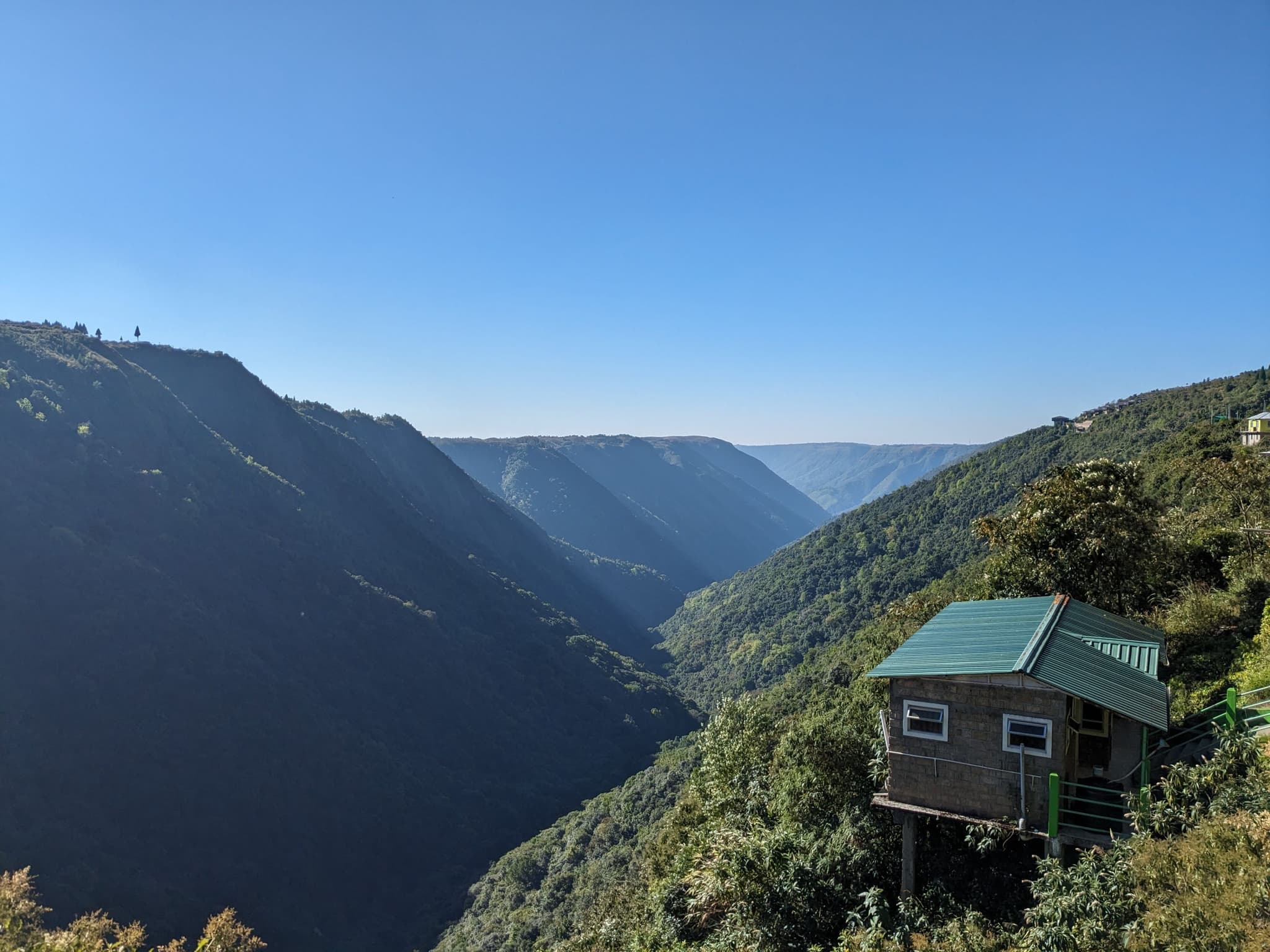 A small wooden cabin with a green roof perched on the edge of a lush, green mountainside, overlooking a deep, expansive valley under a clear blue sky