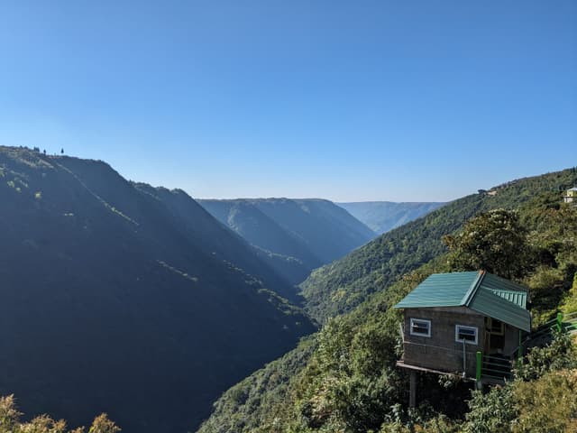 A small wooden cabin with a green roof perched on the edge of a lush, green mountainside, overlooking a deep, expansive valley under a clear blue sky