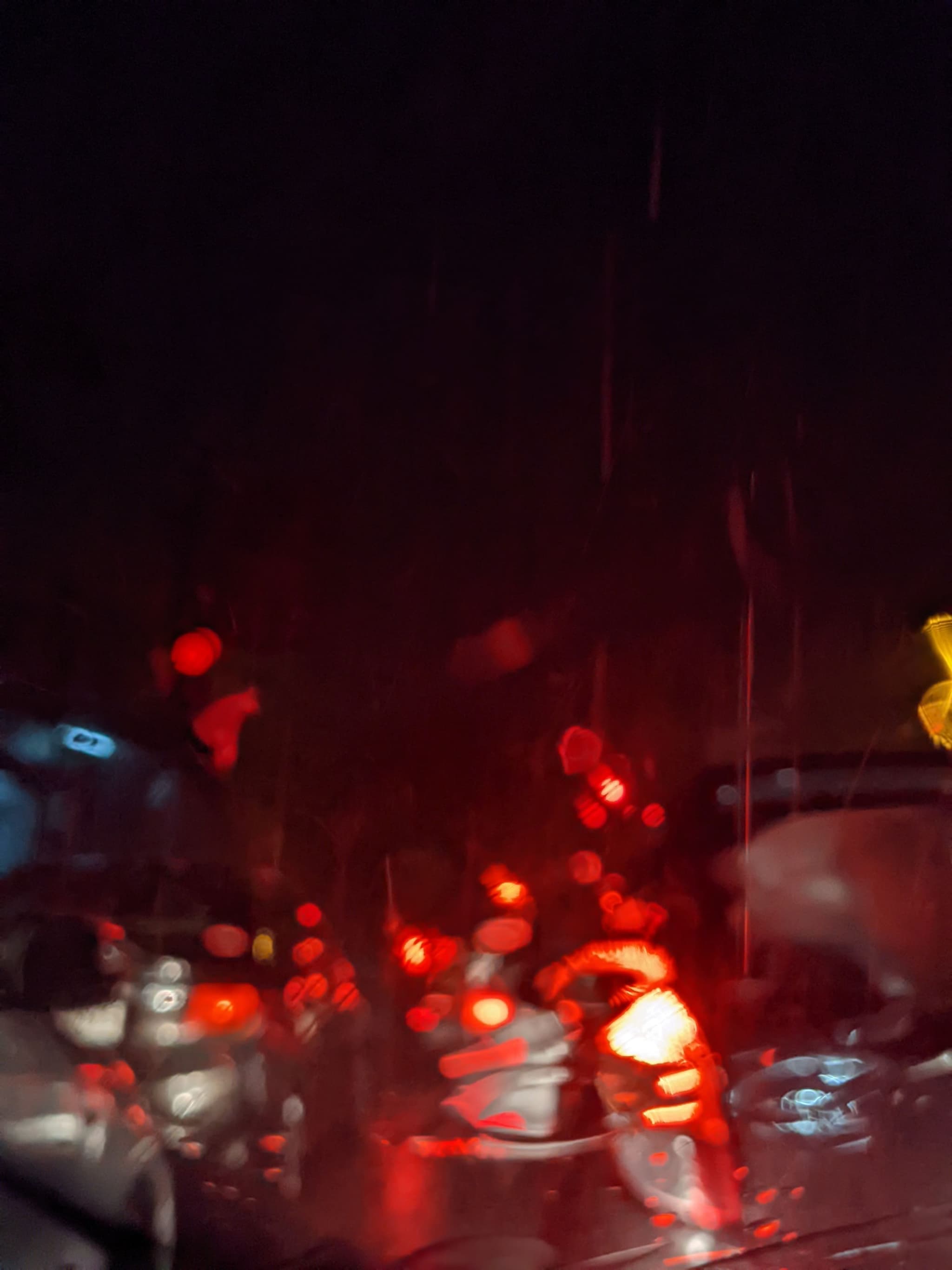 Nighttime traffic viewed through a rain-soaked windshield, with blurred red brake lights and reflections creating a vibrant, abstract scene