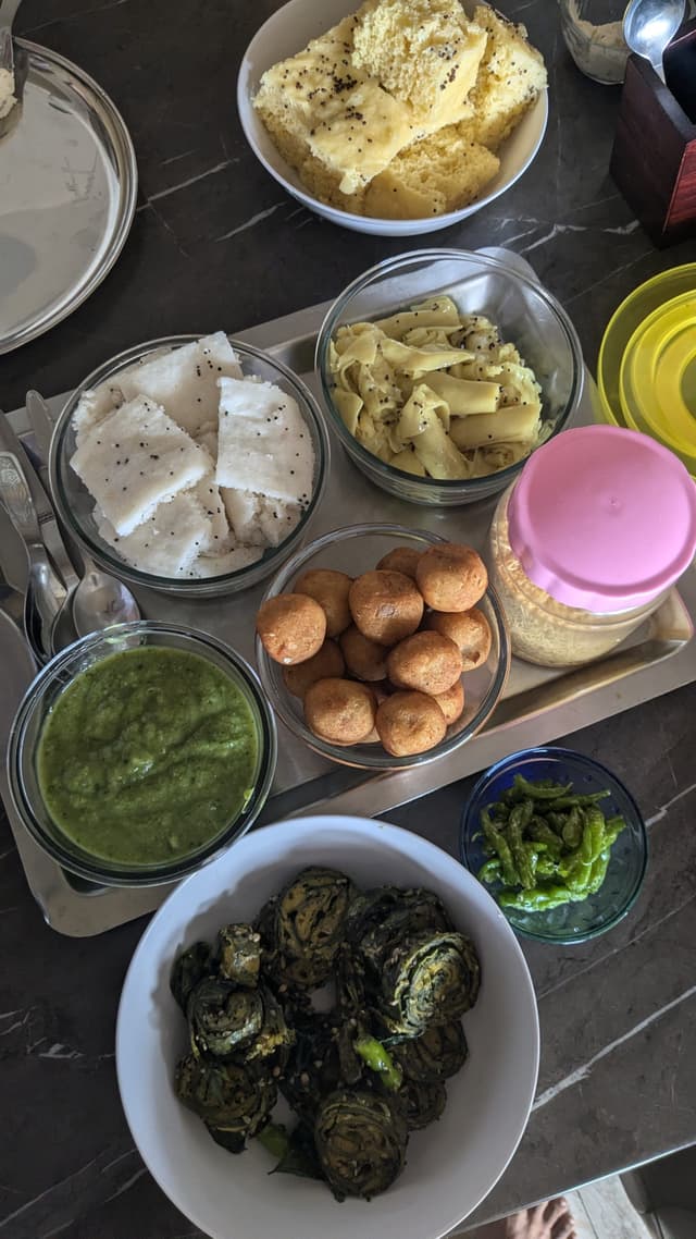 A variety of Indian breakfast foods including chutney, steamed idlis, fried snacks, sliced vegetables, and curry on a table