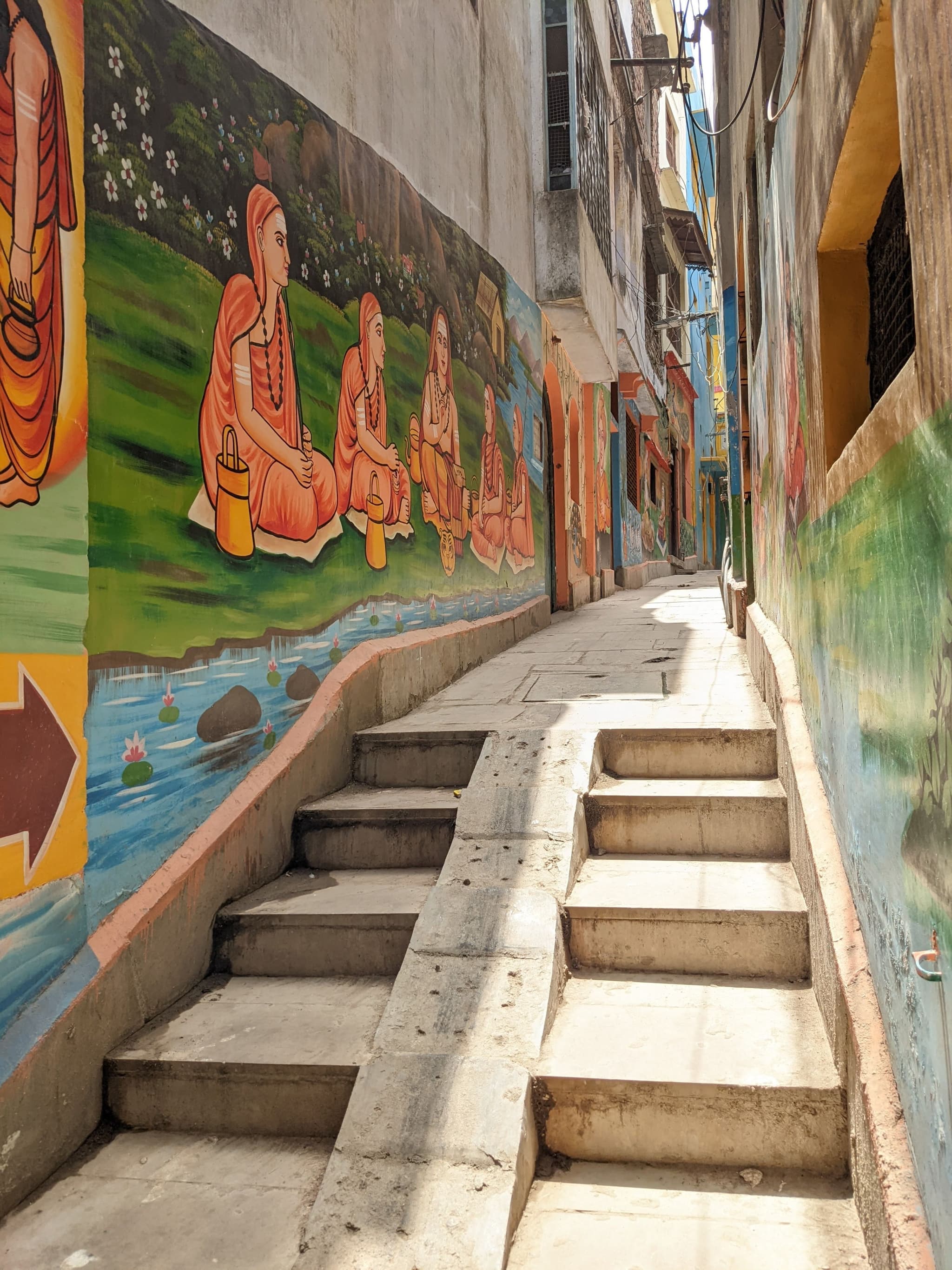 A narrow alley with stair steps painted with murals depicting scenes of people meditating in a natural setting. Sunlight illuminates the path, creating a warm atmosphere in the urban environment