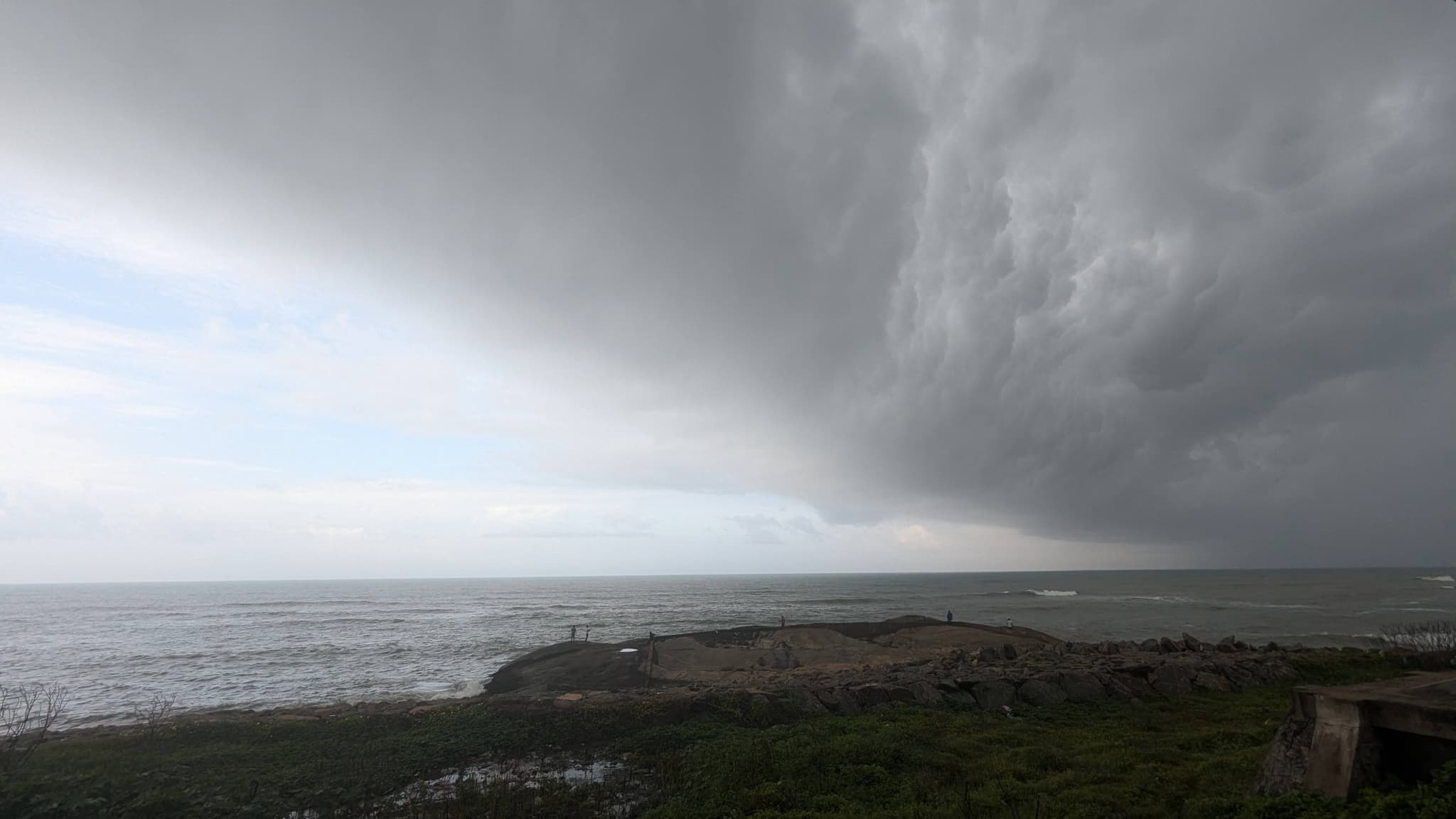 Dramatic storm front with dark clouds sweeping over a coastal shoreline, with ocean waves and grassy dunes in the foreground under a dim, overcast sky