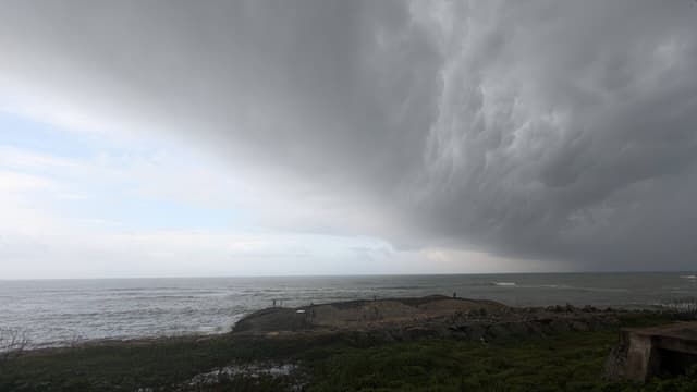 Dramatic storm front with dark clouds sweeping over a coastal shoreline, with ocean waves and grassy dunes in the foreground under a dim, overcast sky
