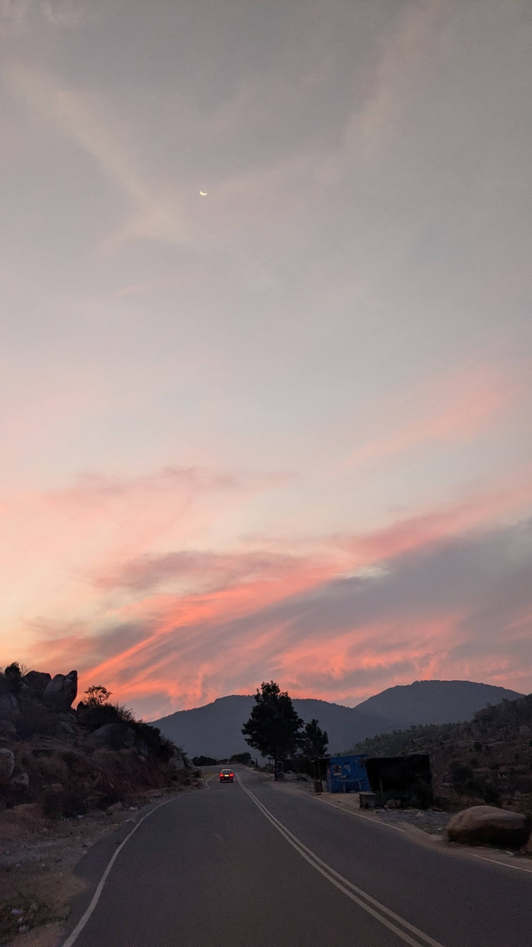 Empty paved road curving through rocky terrain toward distant hills under a pastel pink sunset sky