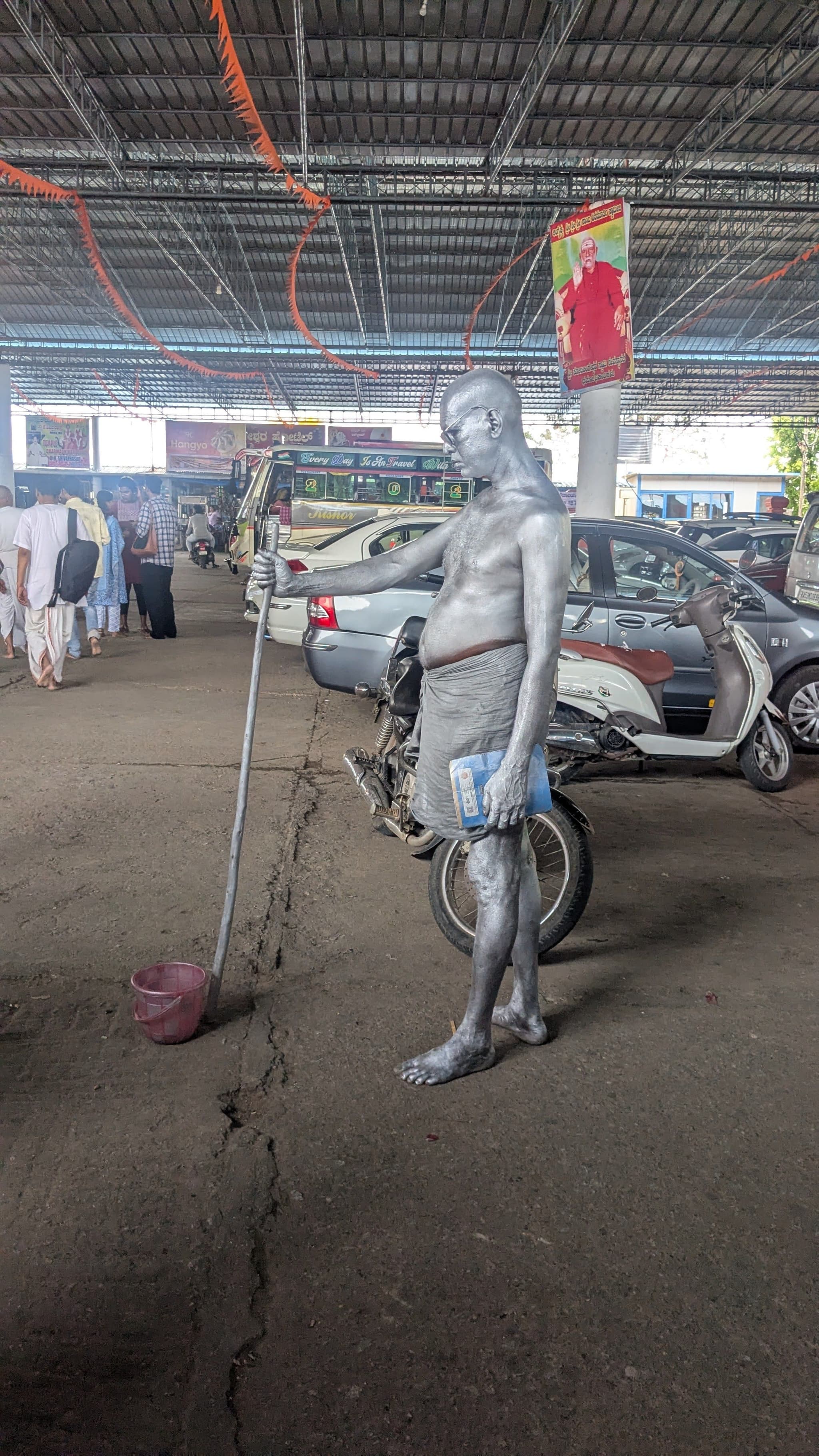 A silver-painted mannequin stands holding a stick in a covered parking area with cars and people in the background