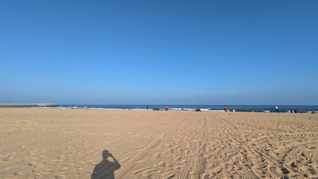 Wide sandy beach under a clear blue sky with the ocean on the horizon and a few distant beachgoers; a person’s shadow in the foreground